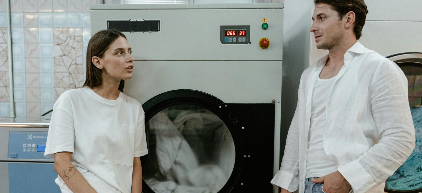 Two adults in a modern launderette having a casual conversation by washing machines.